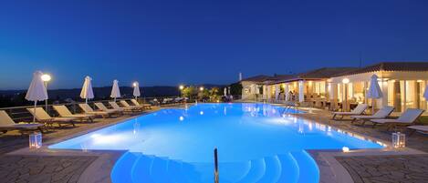 Une piscine entourée de chaises longues et de parasols, le tout bordé de dalles de pierre et adjacent à un bâtiment illuminé.