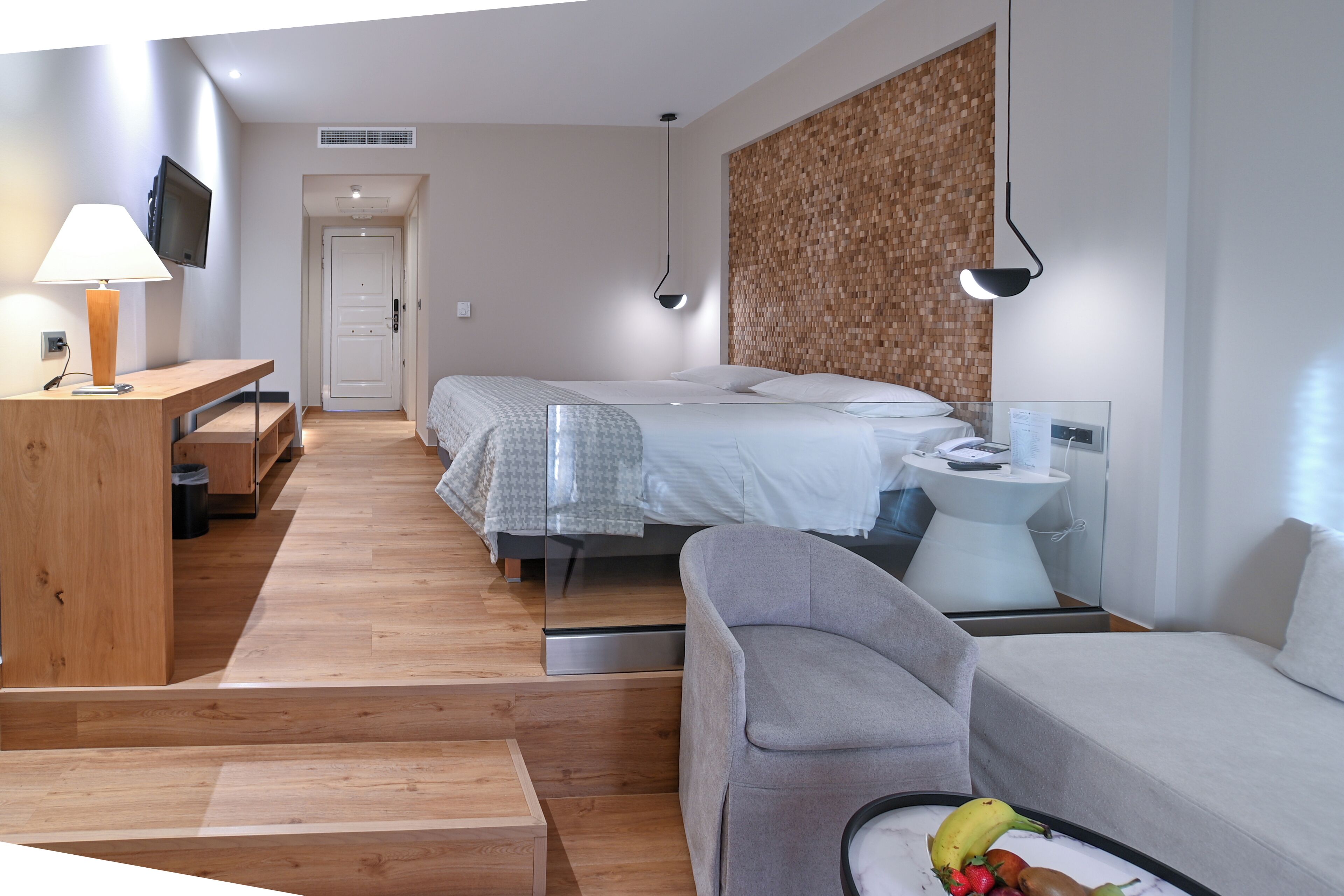 A modern hotel room with two beds, a desk, a sofa, and a small table with a fruit bowl.