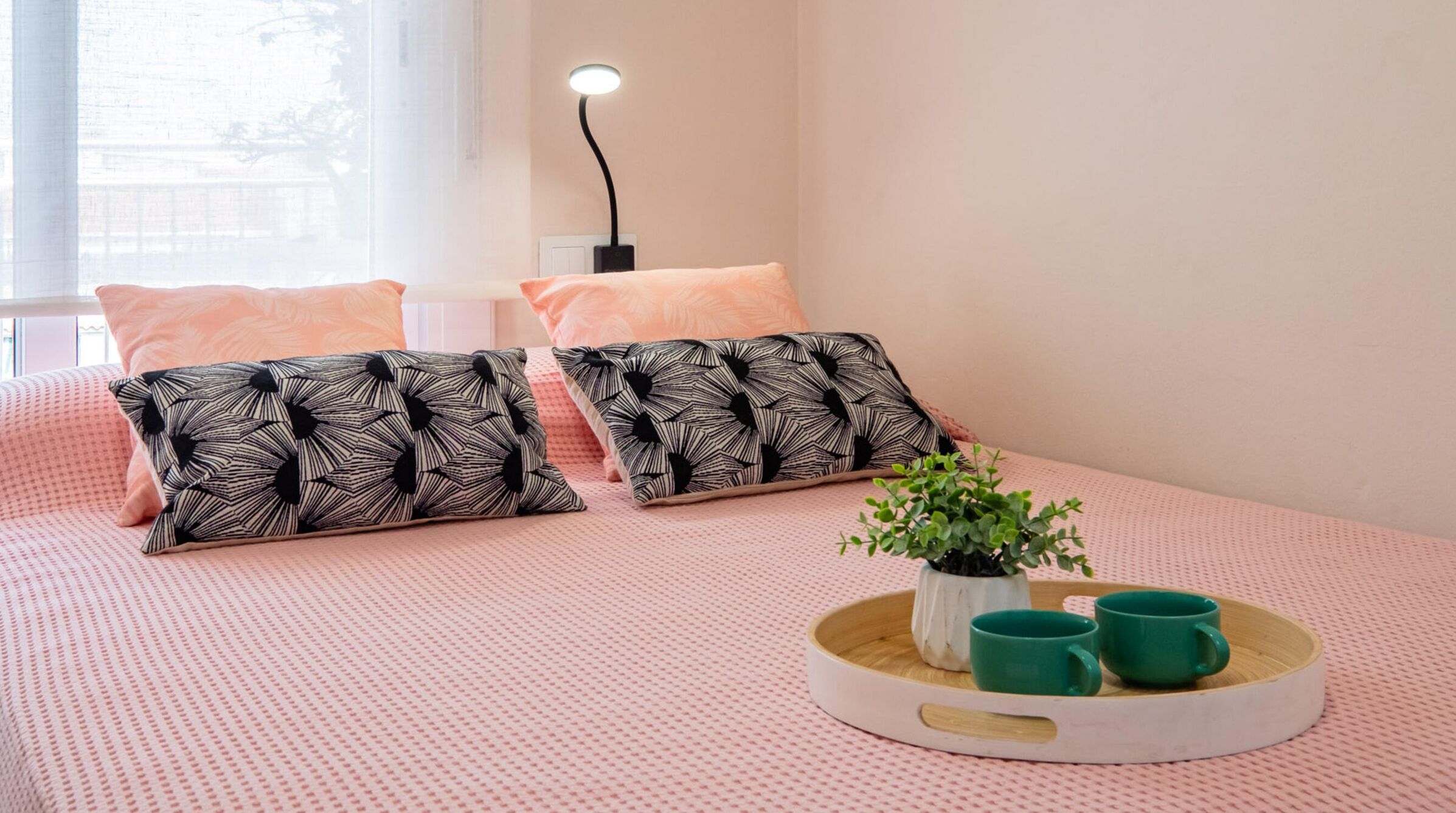 Une chambre à coucher bien rangée, avec une couvre-lit à motifs roses, deux coussins à motifs noir et blanc, une lampe de chevet et un plateau sur lequel se trouvent une plante et deux tasses vertes.