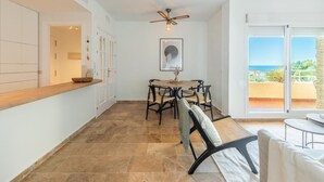 A dining area with a round table and chairs, a ceiling fan, and a view of the sea through large sliding doors.