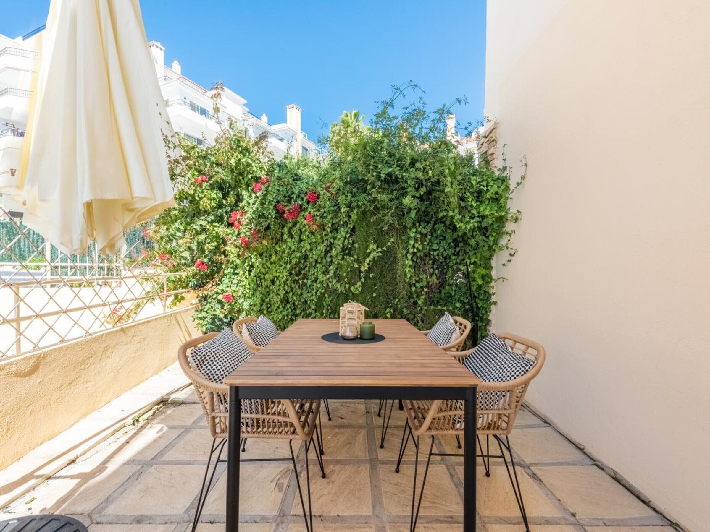 A wooden table with four wicker chairs, a white umbrella, and a wall covered in green foliage and red flowers.