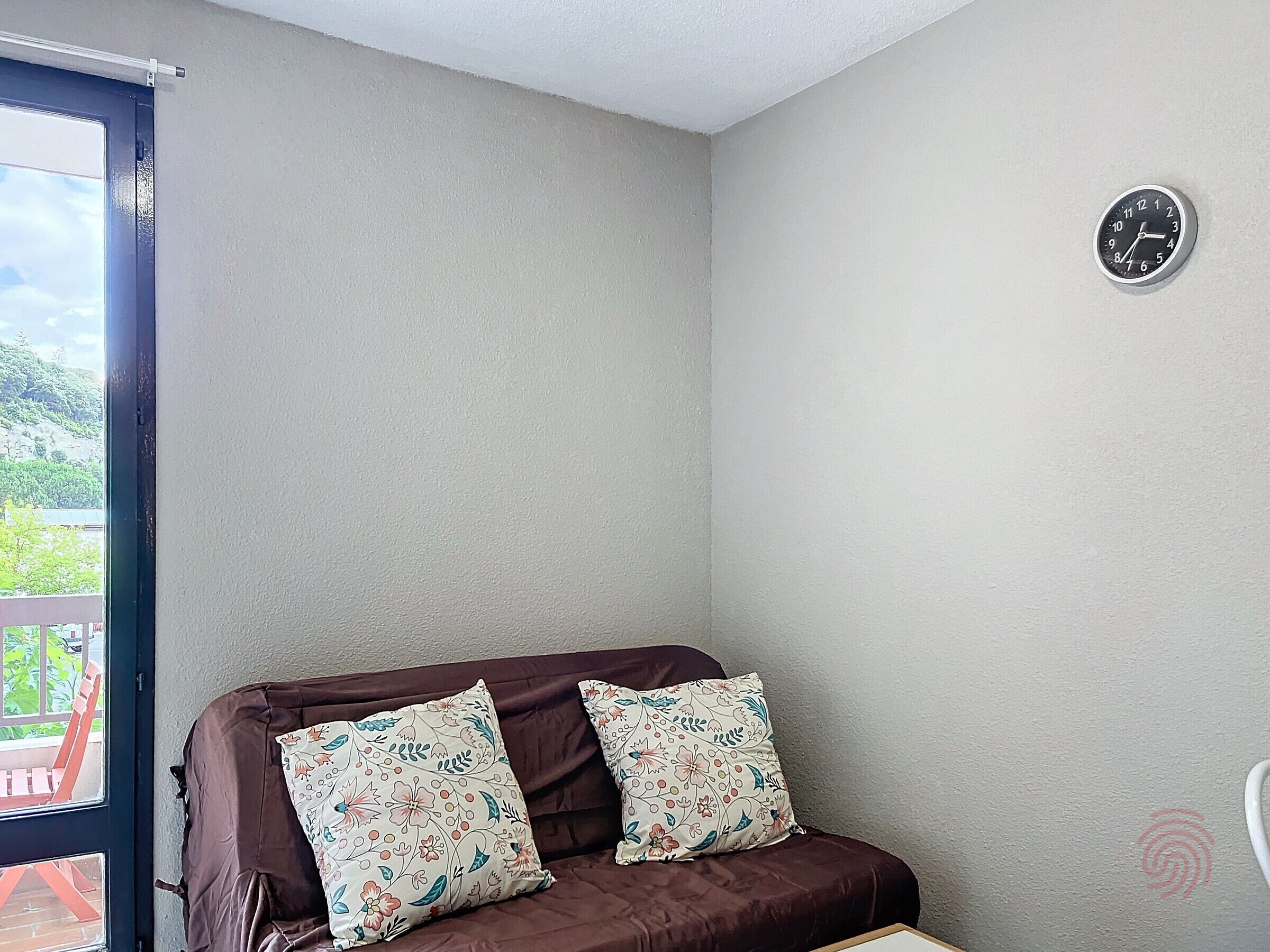 Une pièce avec un canapé marron, deux coussins à motifs floraux, une horloge au mur et une fenêtre donnant sur un paysage verdoyant.