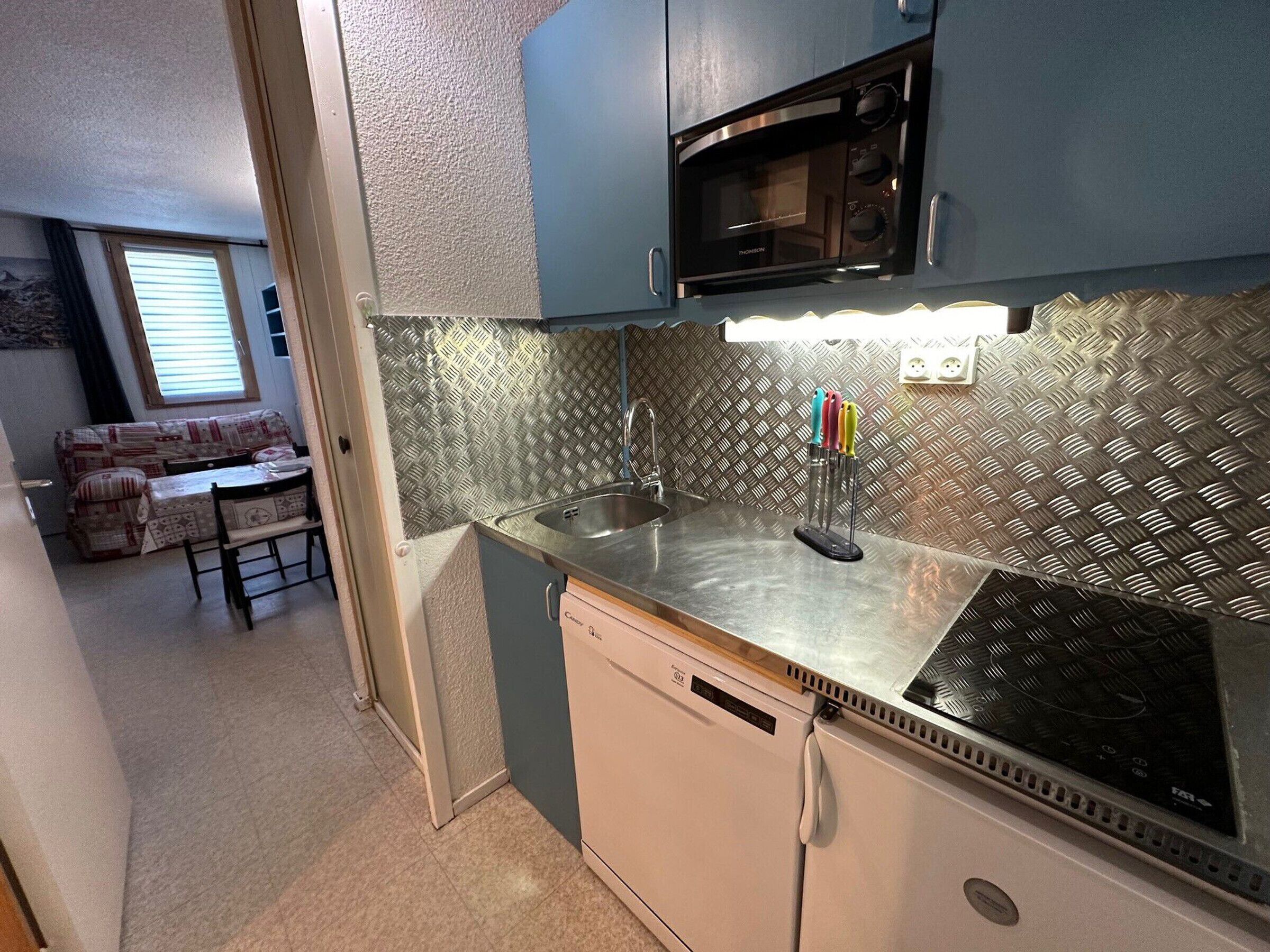 Cocina compacta con microondas, pileta y lavavajillas. Se ve una zona de comedor con sofá y mesa al fondo.