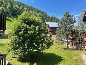 A mountain lodge with a large tree in the foreground, a wooden deck, and a view of the surrounding forest and hills.