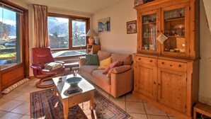 A cozy living room with a red armchair, a beige sofa, a wooden cabinet, and a view of mountains through large windows.
