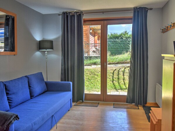 A room with a blue sofa, a floor lamp, and a sliding glass door leading to an outdoor area with a fence and greenery. - Cozy studio in Morzine, near center, 4 pers, terrace (Morzine)