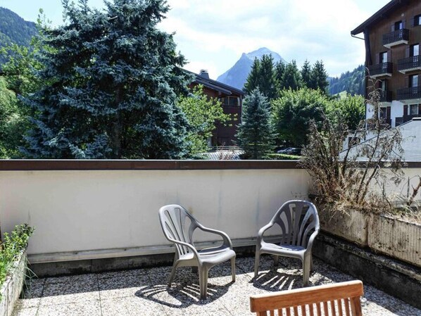 A balcony with two plastic chairs and a wooden bench, overlooking a mountainous landscape with trees and buildings.