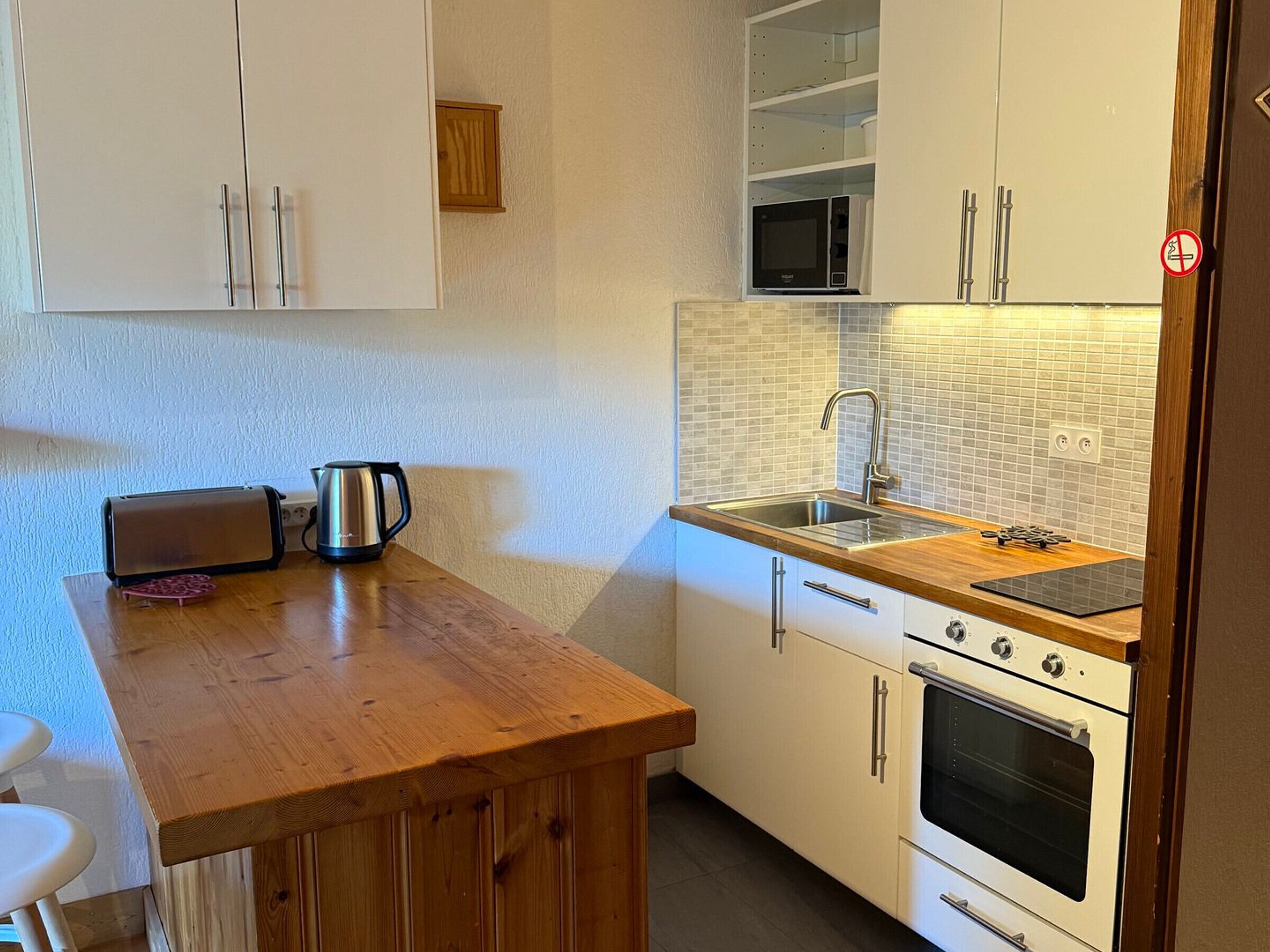 Eine kleine Küche mit weißen Schränken, einer Holz Arbeitsplatte, einer Mikrowelle, einem Wasserkocher und einem Backofen.