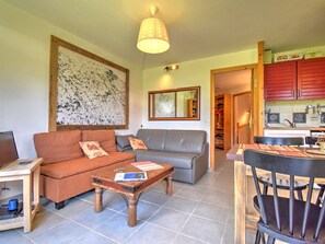 A living room with a sofa, a coffee table, and a dining table with chairs. There is a kitchen area with red cabinets and a sink.