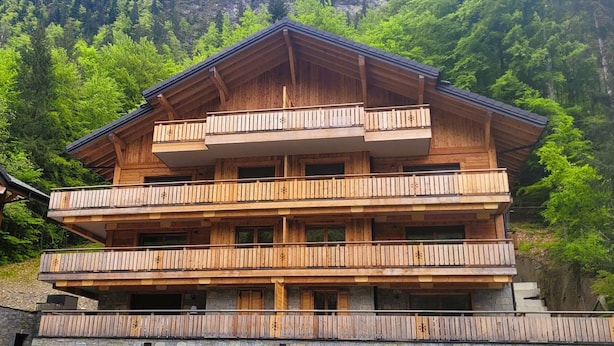 Una cabaña de madera de varios pisos con balcones, con un paisaje de montañas boscosas al fondo.