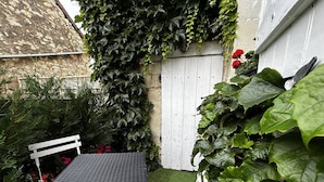 Un petit espace extérieur aménagé avec une table et des chaises, entouré d’une végétation luxuriante et d’une clôture blanche.