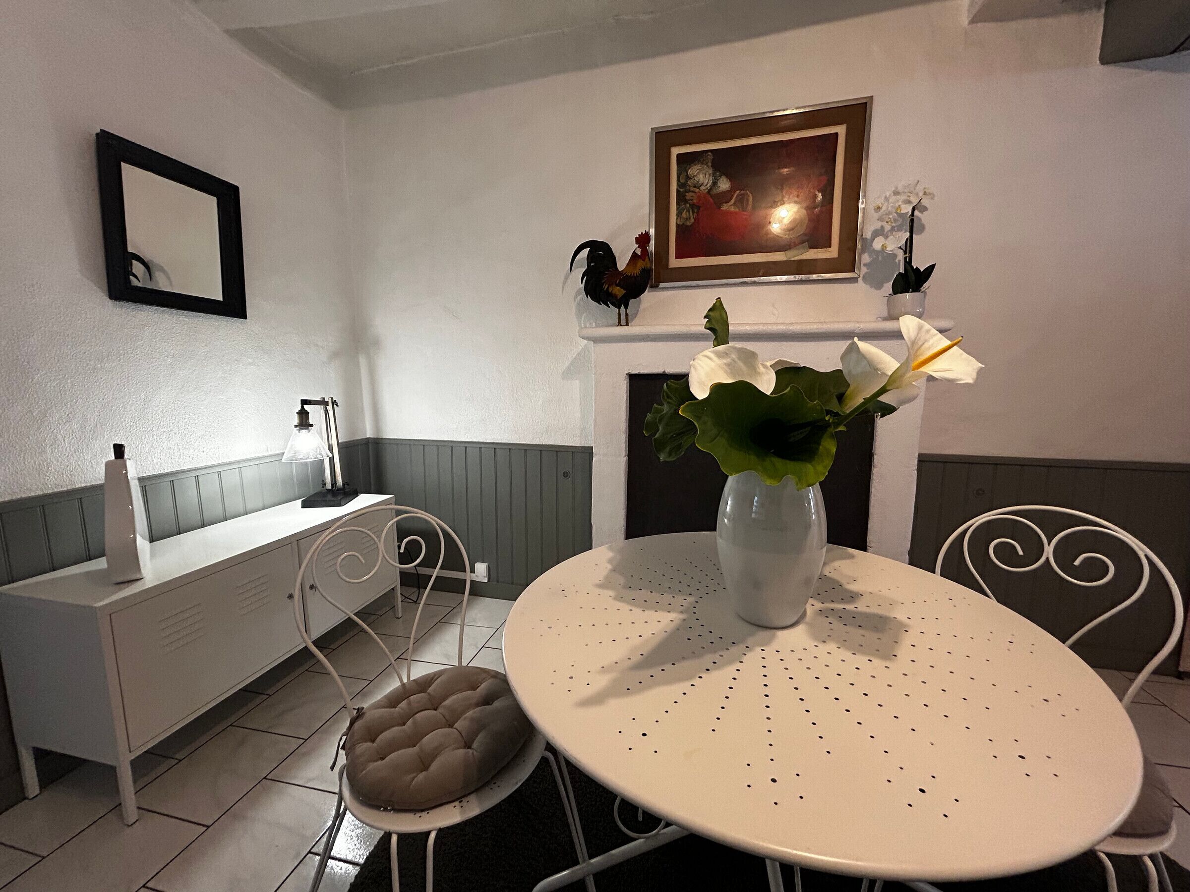Un comedor con una mesa redonda, dos sillas, un banco blanco, un armario blanco, un cuadro enmarcadosobre una chimenea, y un florero con flores blancas en la mesa.