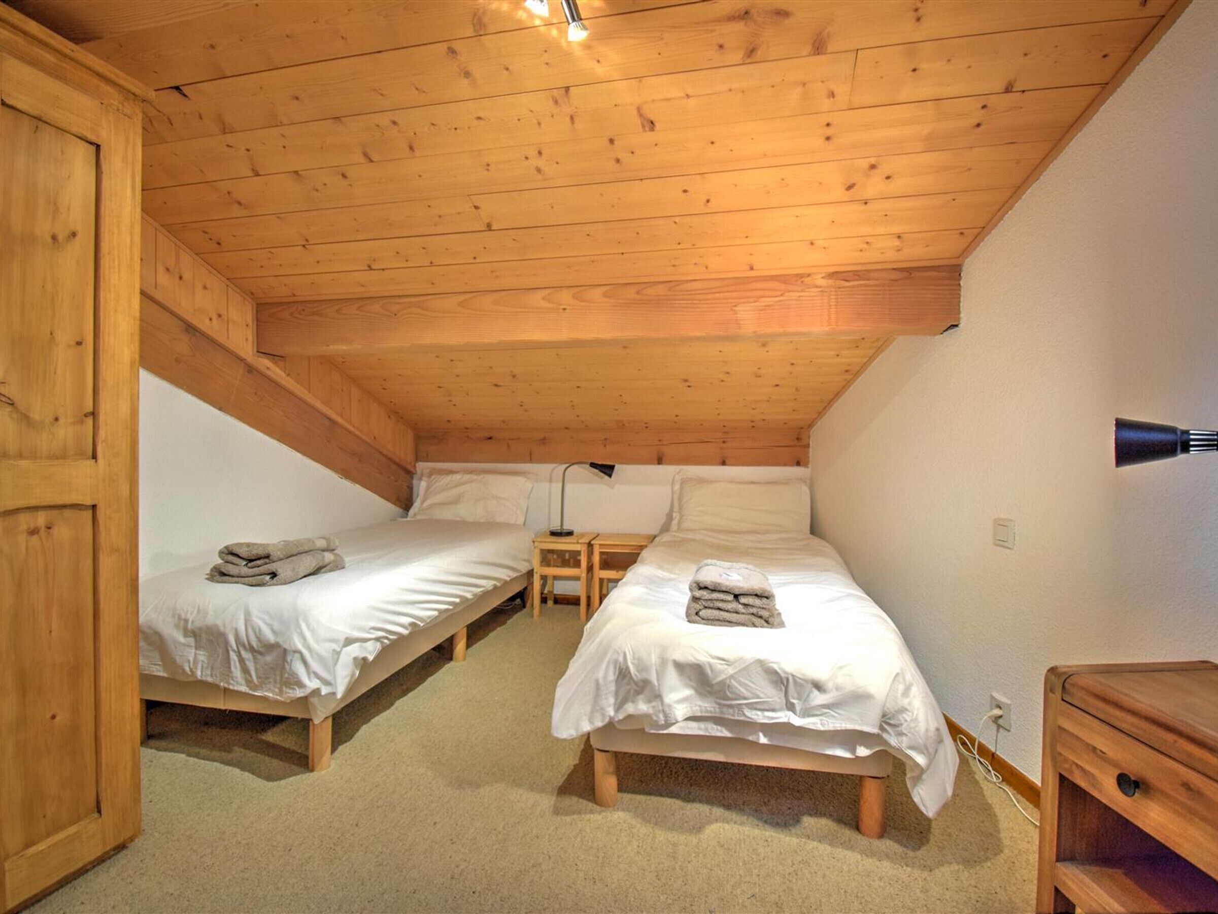 A small, cozy attic bedroom with two single beds, wooden beams on the ceiling, a nightstand, and a lamp.