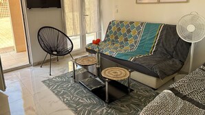 A living room with a sofa, a chair, a small table, a fan, and a TV on the wall. - Cozy 2-Room Apartment with Pool, Tennis, and Parking Near Beach - Sleeps 6 (Fleury-Saint-Pierre-la-Mer)
