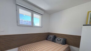 A small, single-bed room with a checkered bedspread, a window with a view of buildings, and a white wall with a wooden headboard. - Cozy 2-Room Apartment with Pool, Tennis, and Parking Near Beach - Sleeps 6 (Fleury-Saint-Pierre-la-Mer)