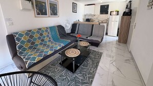 A modern living room with a sofa, a small table, and a chair. There is a kitchen area in the background with a refrigerator and a microwave. - Cozy 2-Room Apartment with Pool, Tennis, and Parking Near Beach - Sleeps 6 (Fleury-Saint-Pierre-la-Mer)