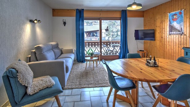 Ein gemütliches Wohnzimmer mit einer Couch, einem Esstisch und Stühlen. An der Wand ist ein Fernseher angebracht, die Decke ist aus Holz und es gibt ein Fenster mit Vorhängen.