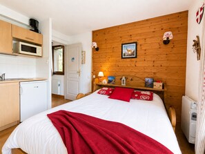 A bedroom with a wooden headboard, a bed with white bedding and red pillows, a bedside table with a lamp, and a view of a kitchen area with a microwave.