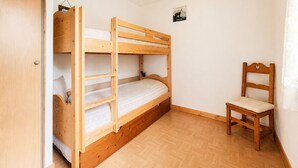 Un lit superposé avec une structure en bois et une chaise en bois, dans une pièce aux murs blancs et au plancher en bois.
