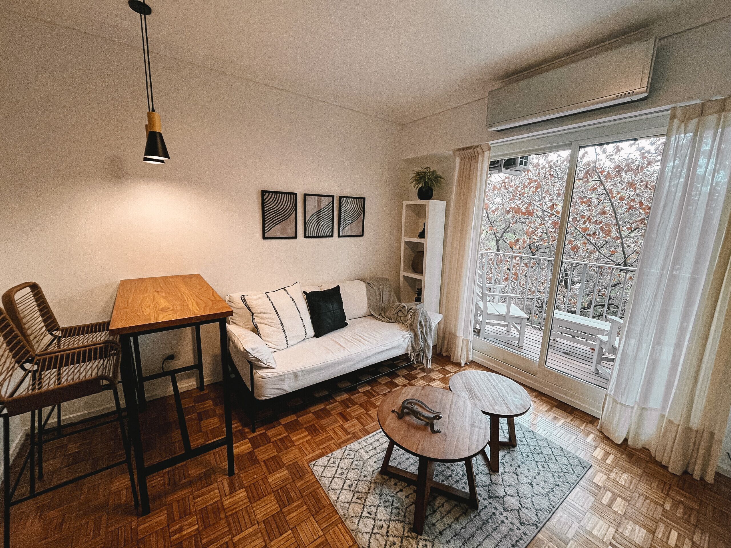 Ein Wohnzimmer mit einer Couch, einem Holztisch, einem Couchtisch, einer Pendelleuchte, einem Bücherregal und einem großen Fenster mit Blick auf Bäume.