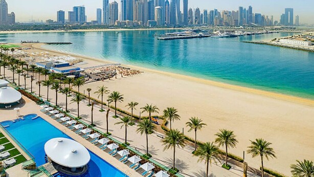 Un complejo turístico frente al mar con piscina, playa y el horizonte de la ciudad al fondo.