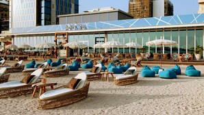Un área de descanso en la playa con sillones de mimbre y pufs azules, un edificio de vidrio con letrero 'Roto Bay' y sombrillas.