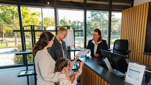 A reception desk with a computer, a whiteboard, and a window with a view of trees and a car. - Wooden Lodge Near Three National Parks (Ruinen)