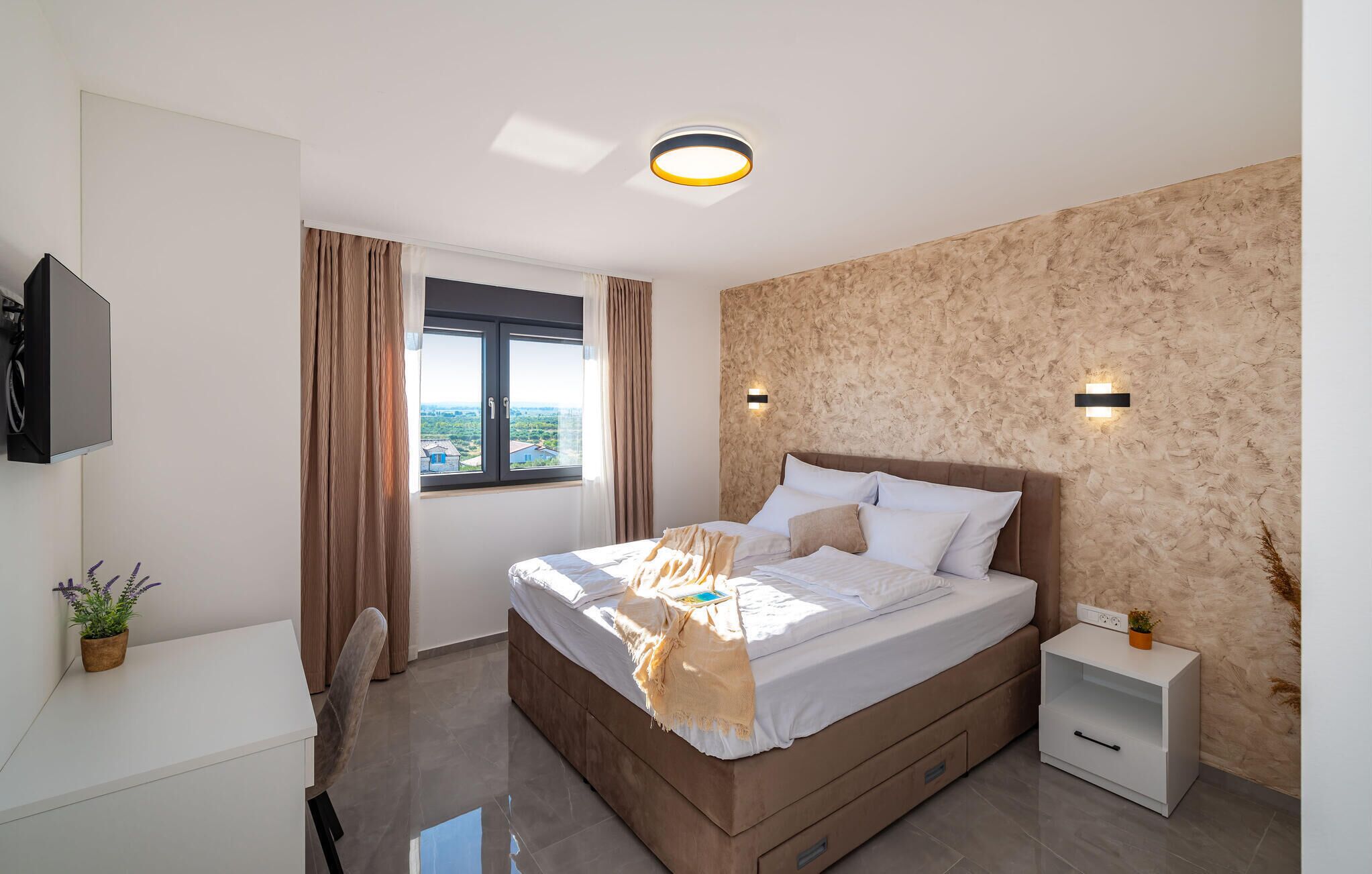 A modern bedroom with a large bed, a TV mounted on the wall, a window with curtains, and a textured wall.
