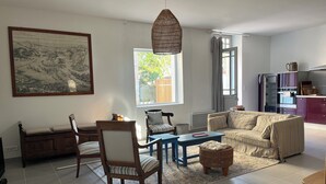 Ein Wohnzimmer mit einer Couch, Sesseln, einem Couchtisch, einem Esstisch, einem großen Gemälde, einer Hängelampe und einer Küchenzeile mit Schränken und Geräten.