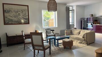 Un salon comprenant un canapé, des fauteuils, une table basse, une table à manger, un grand tableau, un luminaire suspendu et un coin cuisine avec des placards et des appareils électroménagers.