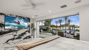 Une chambre dotée d’un grand lit, d’un ventilateur de plafond, d’un téléviseur diffusant des images de surfeurs, et offrant une vue sur un patio extérieur aménagé avec des sièges et des plantes.