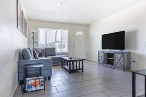 A living room with a grey sofa, a coffee table, a TV on a wooden stand, and a magazine rack. - Cozy 3BR House Near Attractions & Shopping (Phoenix)