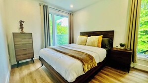 Un dormitorio con cama, cómoda, mesita de noche y ventana con vista a follaje.