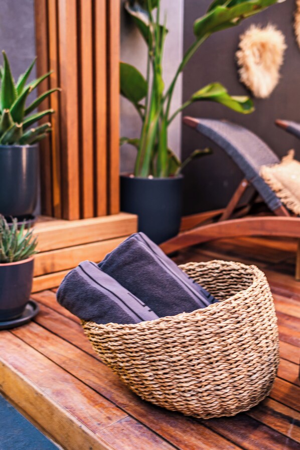 Un panier en osier avec des coussins, posé sur une terrasse en bois, entouré de plantes en pot et d’un mur fait de lattes de bois.
