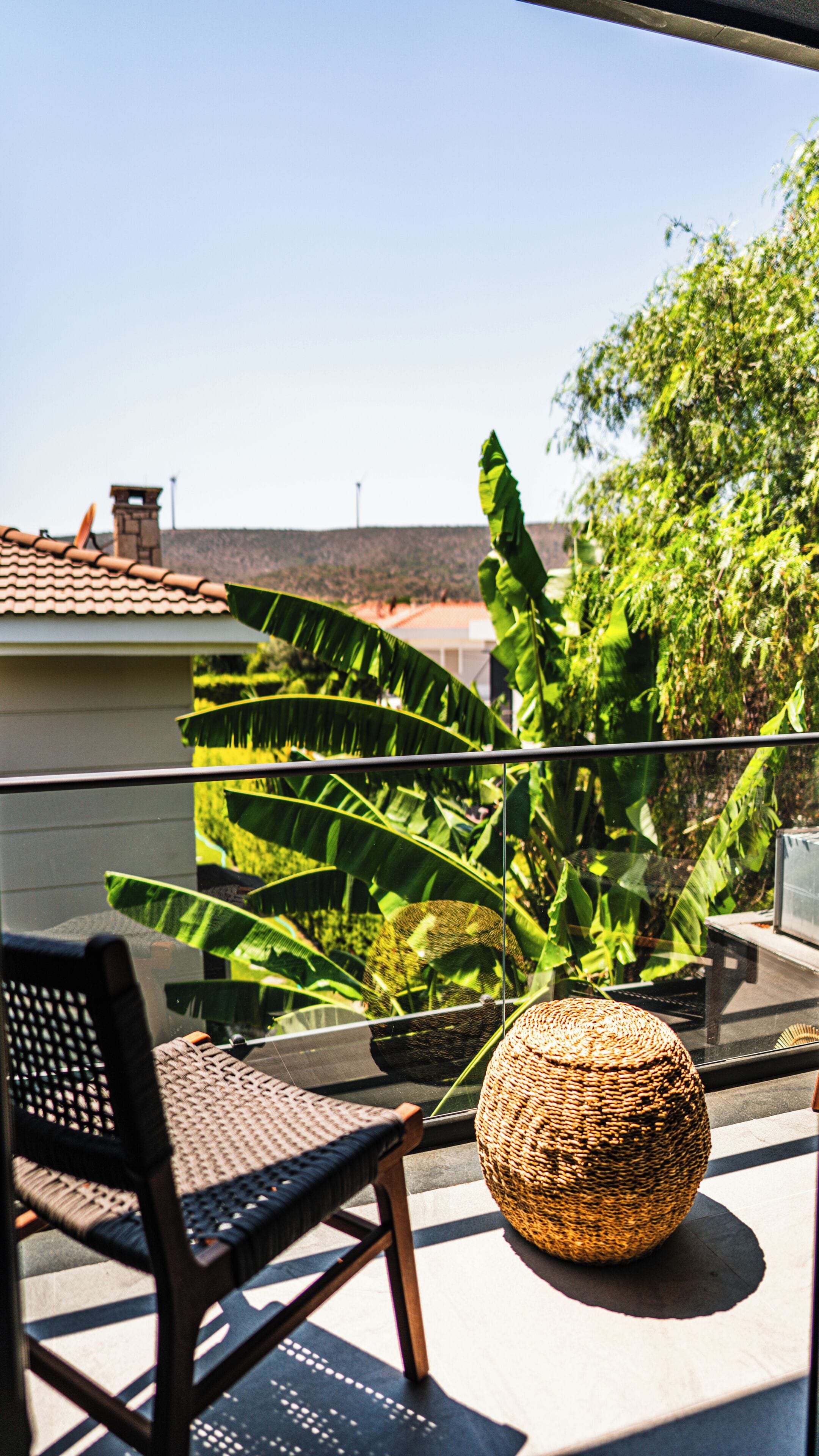 A balcony with a wicker chair and a woven basket, overlooking a garden with large green leaves and a building with a tiled roof.