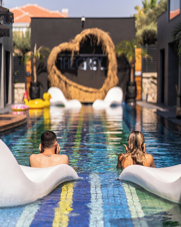 A swimming pool with two people in the water, a heart-shaped structure in the background, and a building with a red roof. - The Grey Alaçatı (Cesme)