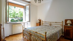 A bedroom with a bed, a window, a radiator, a wooden nightstand, a lamp, and a wooden chair. - Lovely apartment in Monteriggioni (Monteriggioni)