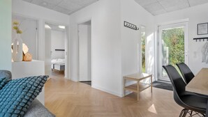 Uma sala de estar moderna e com layout aberto, com piso de madeira em padrão herringbone, uma pequena mesa de madeira e uma cadeira preta. Há uma cama ao fundo, uma porta branca e vista para o verde lá fora.