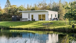 Uma casa moderna com uma grande porta de vidro, um pátio com mobiliário externo e um lago em primeiro plano.