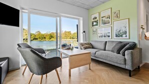 Uma sala de estar moderna com sofá, poltrona e mesa de centro, janelas amplas com vista para um lago e várias fotos emolduradas na parede.