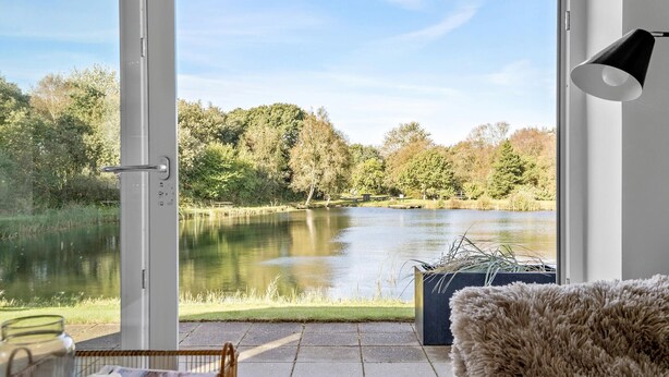 Quarto com uma janela grande com vista para um lago, uma cadeira de vime, um tapete fofo e um abajur preto.