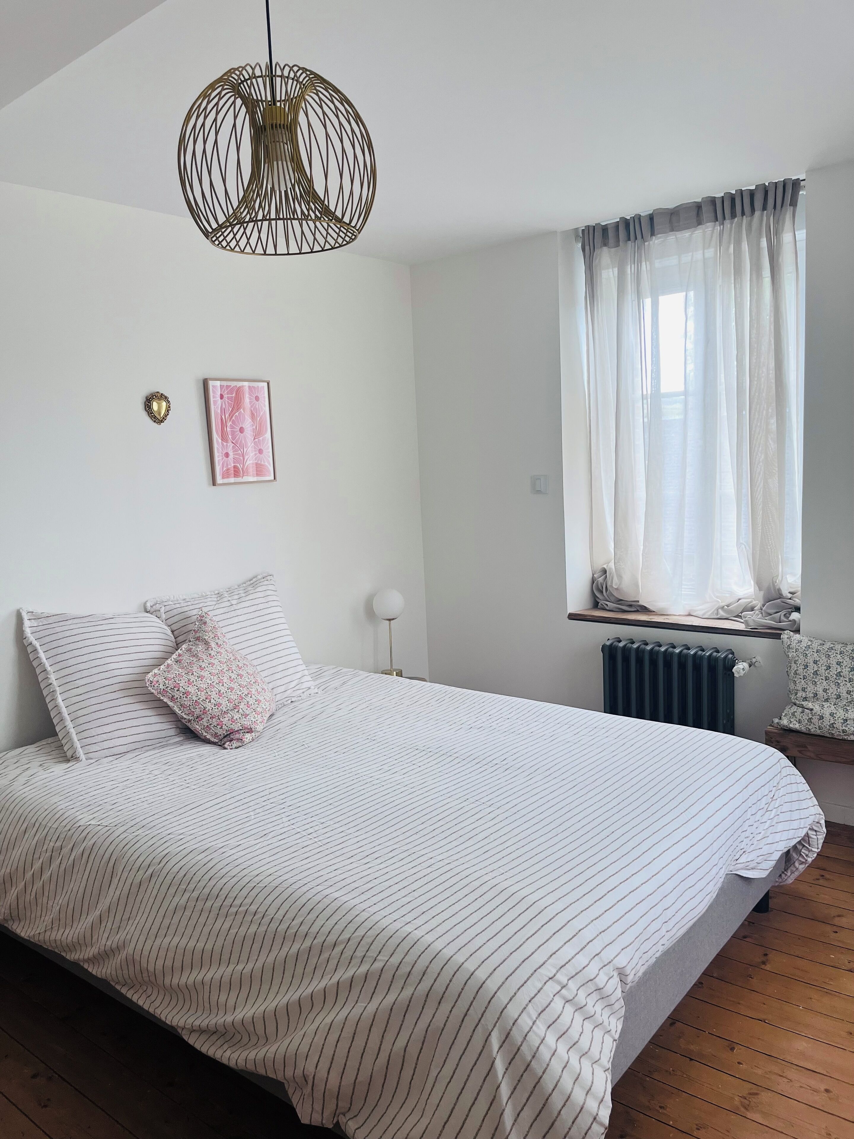 A bedroom with a large bed, a hanging light fixture, a window with sheer curtains, and a radiator.