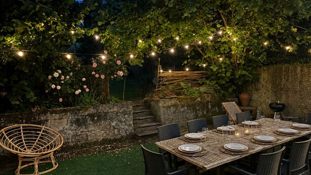 An outdoor dining area with a table set for four, surrounded by chairs, under string lights and climbing roses.