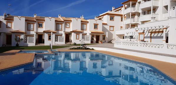 A pool area with a clear blue pool, surrounded by white buildings with balconies and a tiled roof.