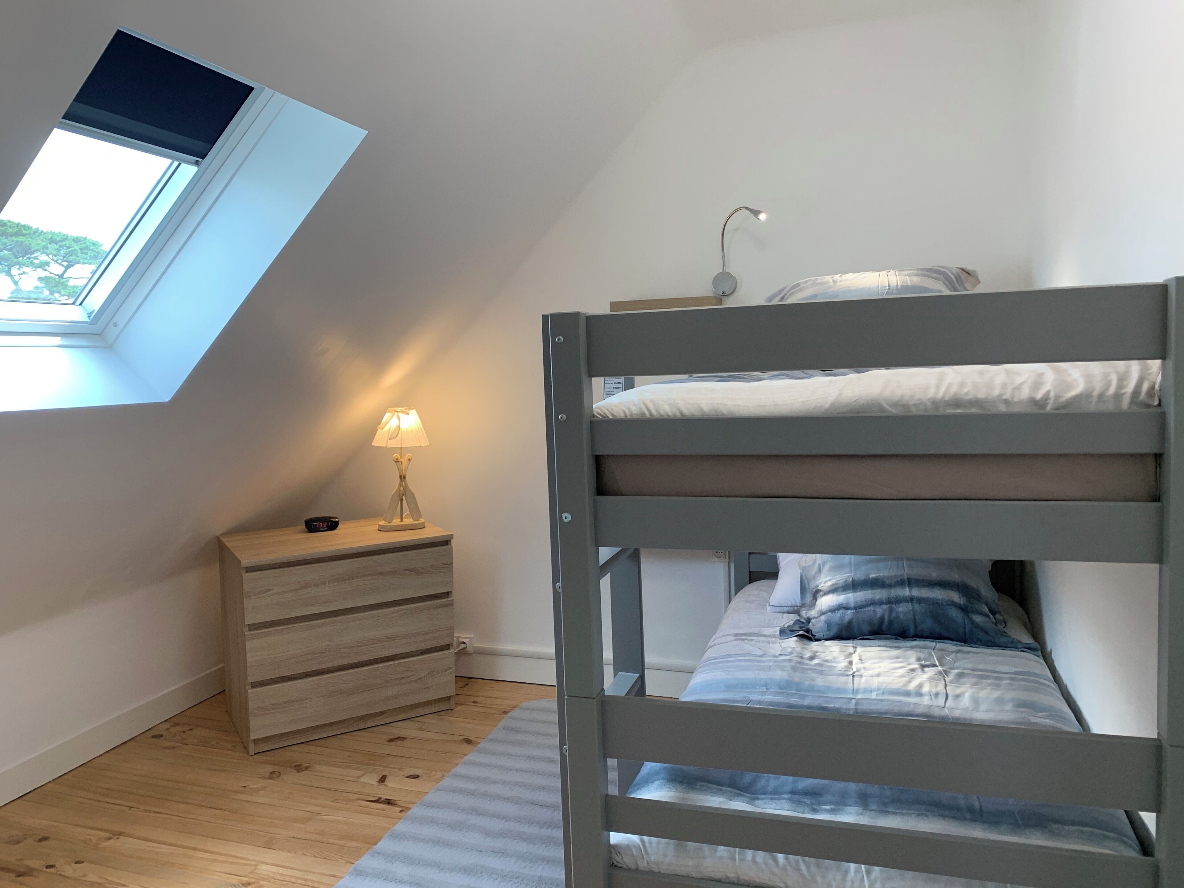 A bedroom with a bunk bed, a bedside table with a lamp, a wooden dresser, and a skylight.