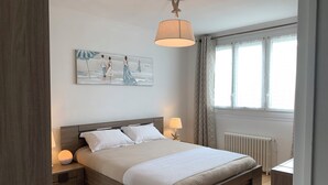 Quarto com cama de madeira, roupa de cama branca, abajur em formato de estrela-do-mar, pintura na parede e janela com cortinas.