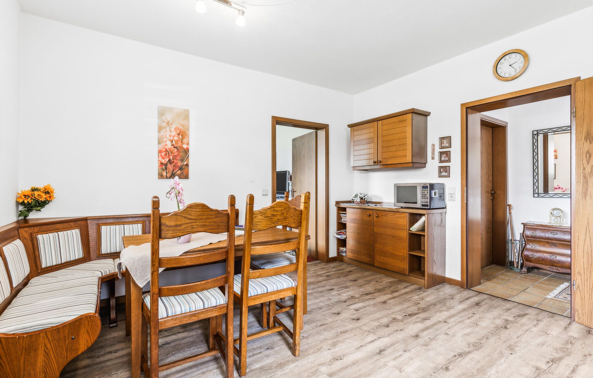 Ein Zimmer mit einem Holzesstisch, Stühlen, einer Mikrowelle, einem Schrank und einer Uhr an der Wand.
