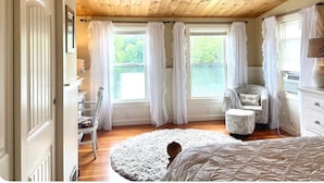 Un dormitorio con techo de madera, ventanas amplias con cortinas blancas, una cama, una silla y un aparador.