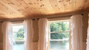 Una habitación con pisos de madera, tres ventanas con cortinas blancas y vistas a un lago.