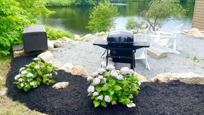 Un patio trasero con una parrilla, sillas de exterior y vistas a un lago rodeado de árboles.
